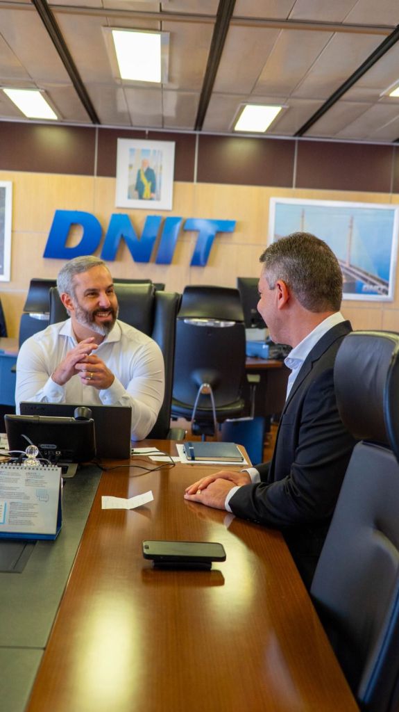 Reunião no DNIT entre dois homens discutindo sobre a triplicação da BR-230, com mesas equipadas em um ambiente de escritório.