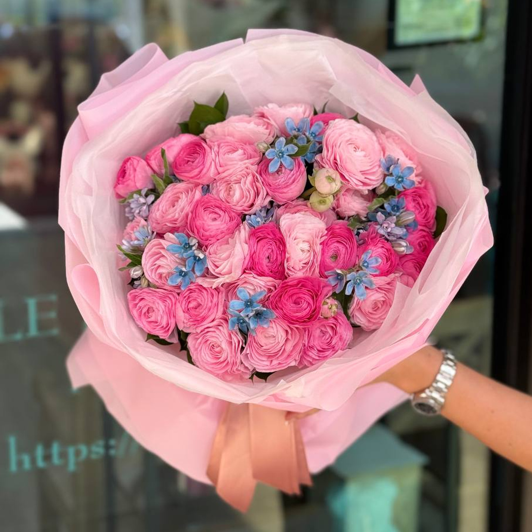 Pink Reverie pink ranunculus bouquet with blue accent flowers, blush pink wrapping, and satin ribbon