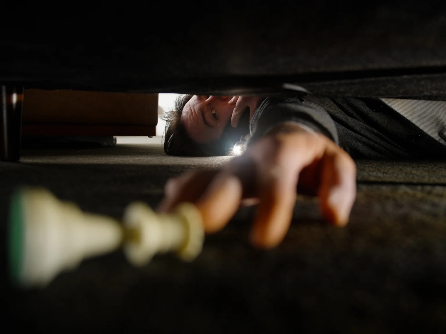 Person crawling under a bed in a dim room, reaching toward the foreground with a flashlight nearby.