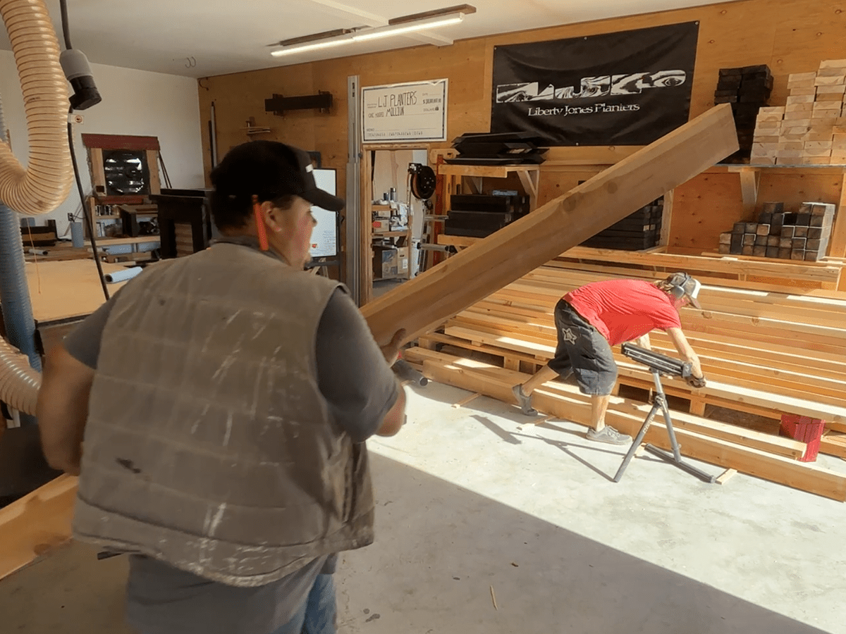 wo craftsmen working in the Liberty Jones Planters woodshop—one carrying a large cedar board, the other positioning lumber on a sawhorse. Stacks of finished wood and a branded banner are visible in the background, capturing the hands-on craftsmanship behind each planter.