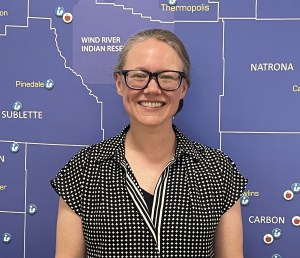 A woman with glasses stands in front of a blue map of Wyoming.