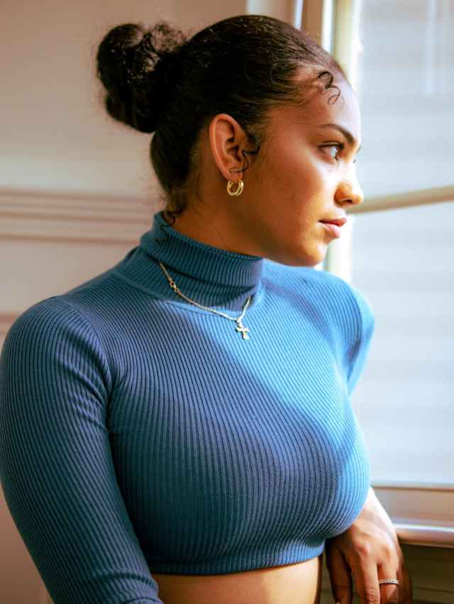 Portrait of a woman in a blue turtleneck gazing out a window, exuding elegance.