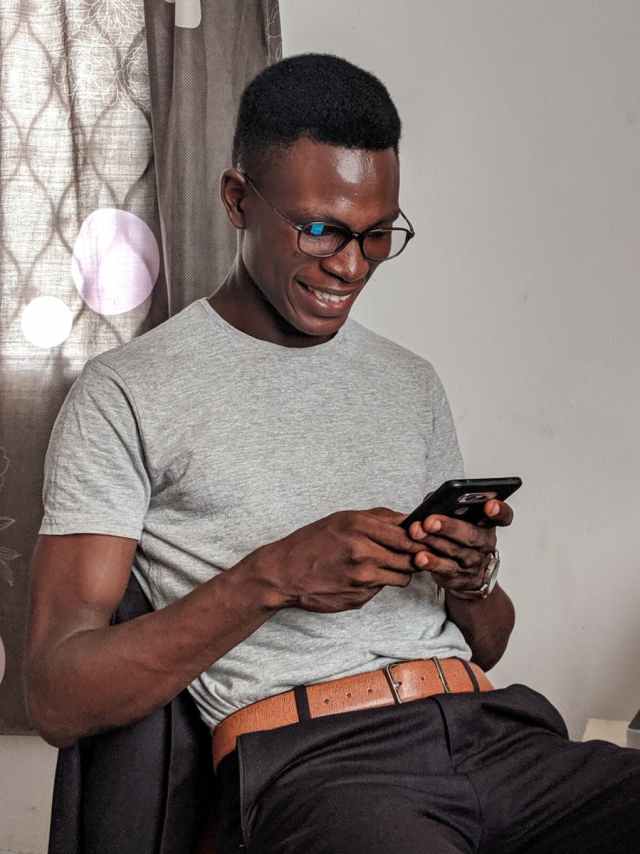 A happy young man uses his smartphone while sitting indoors, expressing joy.