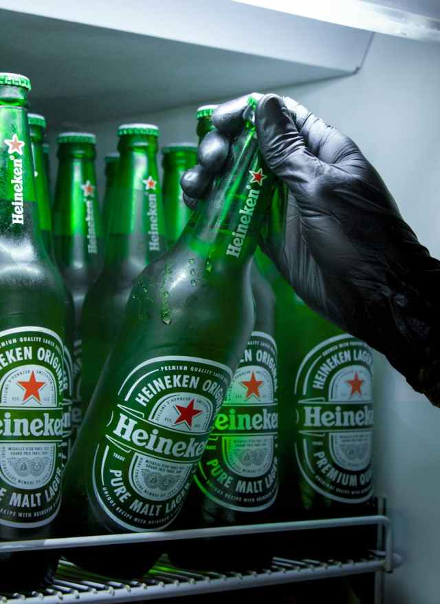 Gloved hand grabbing a Heineken beer bottle from a refrigerator stocked with cold beverages.