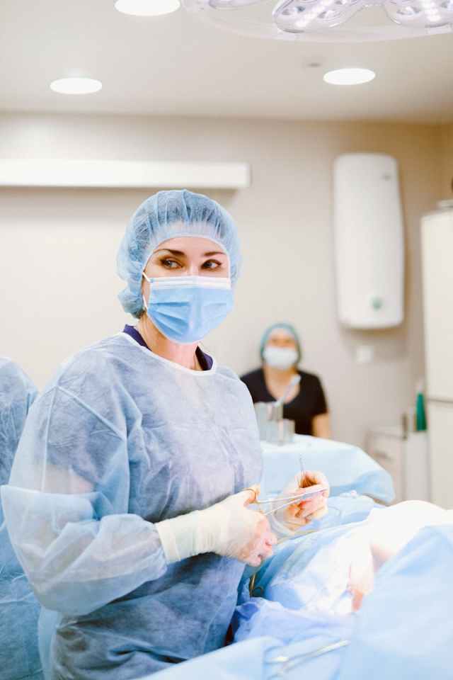 A female surgeon in a blue surgical gown and mask performing surgery in a hospital operating room.