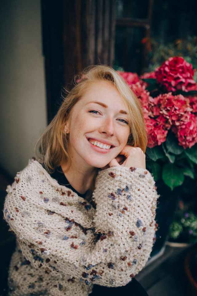 Charming blonde woman smiling in a cozy sweater near vibrant hydrangeas outdoors.