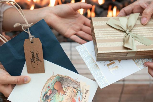 Close-up of hands exchanging beautifully wrapped gifts and thank you cards with a warm fireplace background.
