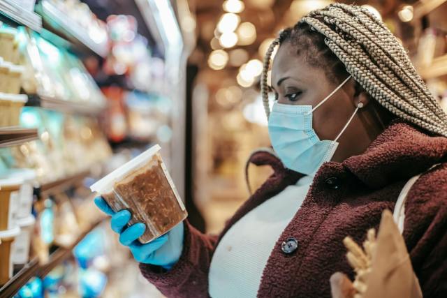 Adult woman in supermarket, choosing groceries with mask and gloves, symbolizes COVID-19 safety.