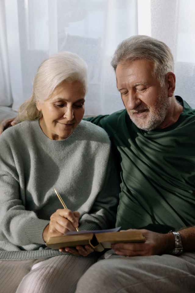 An affectionate elderly couple writing in a notebook while sitting together indoors.
