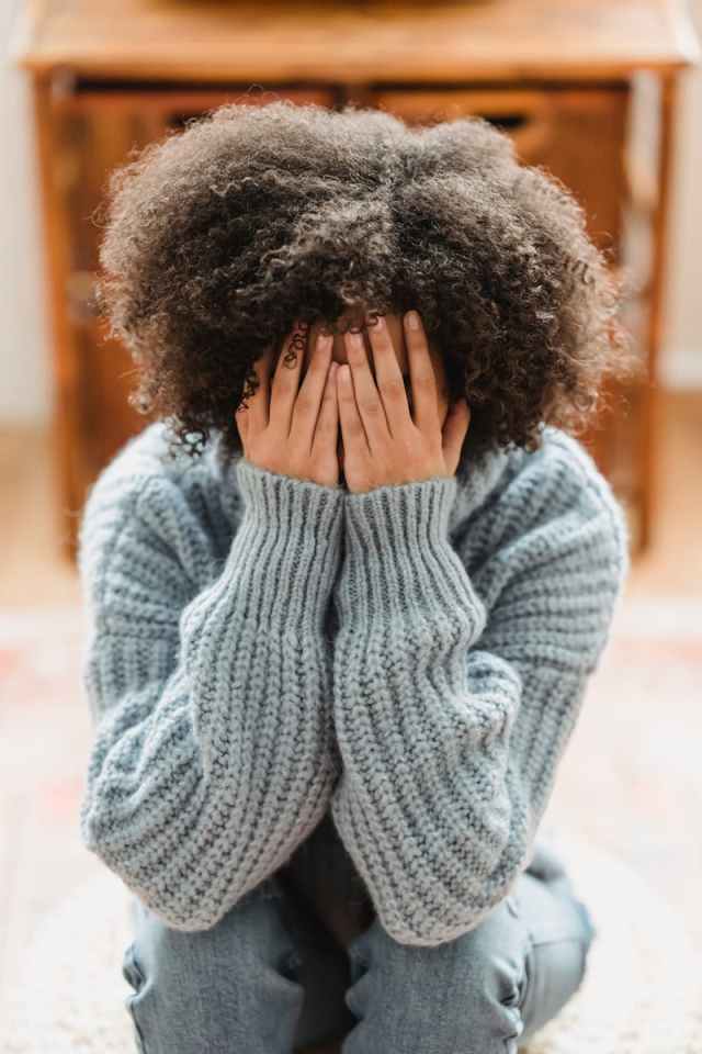 A woman in a knit sweater sits indoors, covering her face with her hands, conveying a sense of emotion.