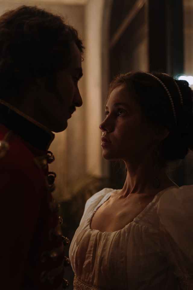A woman and man in historical clothing share an intimate gaze in a dimly lit room.