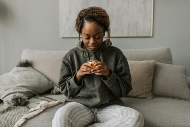 Black woman relaxing on sofa with a cup of tea, creating a cozy stay-at-home vibe.