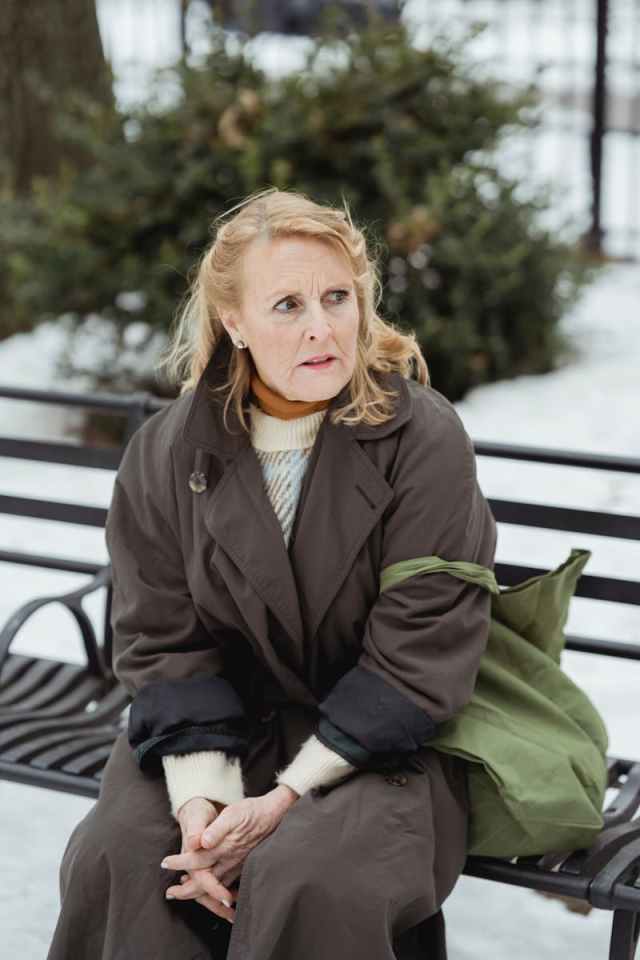 Elderly woman in warm coat sits on a park bench, looking anxious in winter setting.