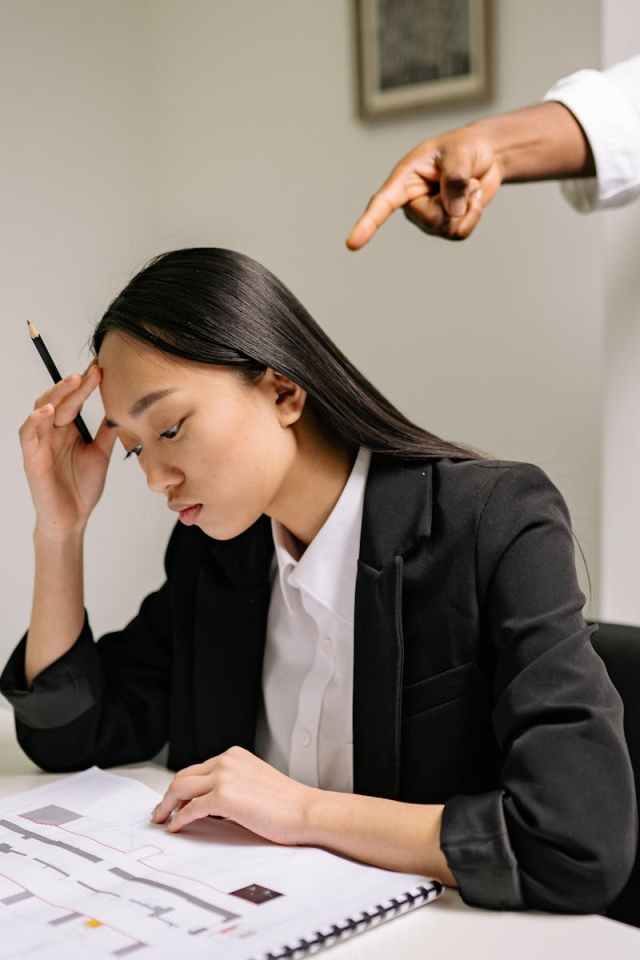 Woman in black blazer studying documents as hand points accusingly, illustrating workplace stress.