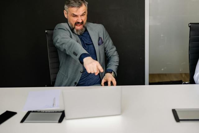 A businessman expressing frustration while pointing at a laptop in a modern office setting.