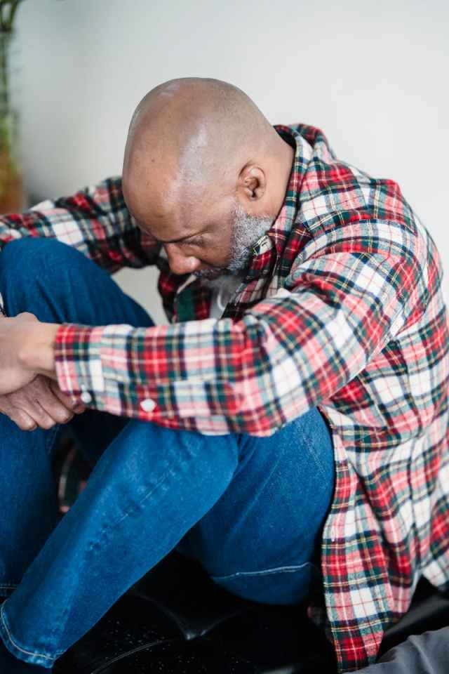 A bald man in a checkered shirt sits indoors, looking contemplative and sad.