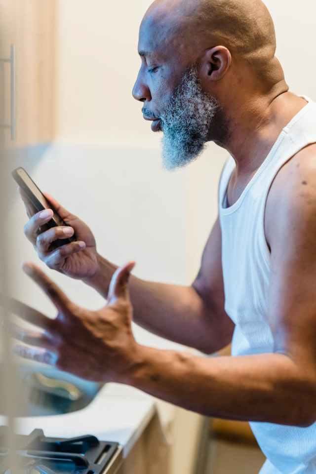 Senior man in kitchen, expressing frustration during a phone call, wearing a white tank top.