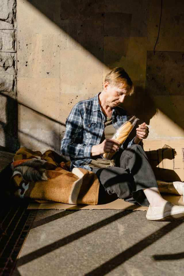 An elderly woman sits on the street in sunlight holding bread, reflecting poverty and homelessness.