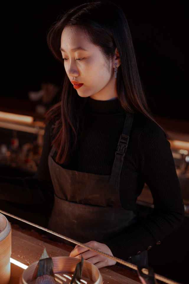 Portrait of a woman with long hair and red lipstick wearing a black apron in a dimly lit setting.
