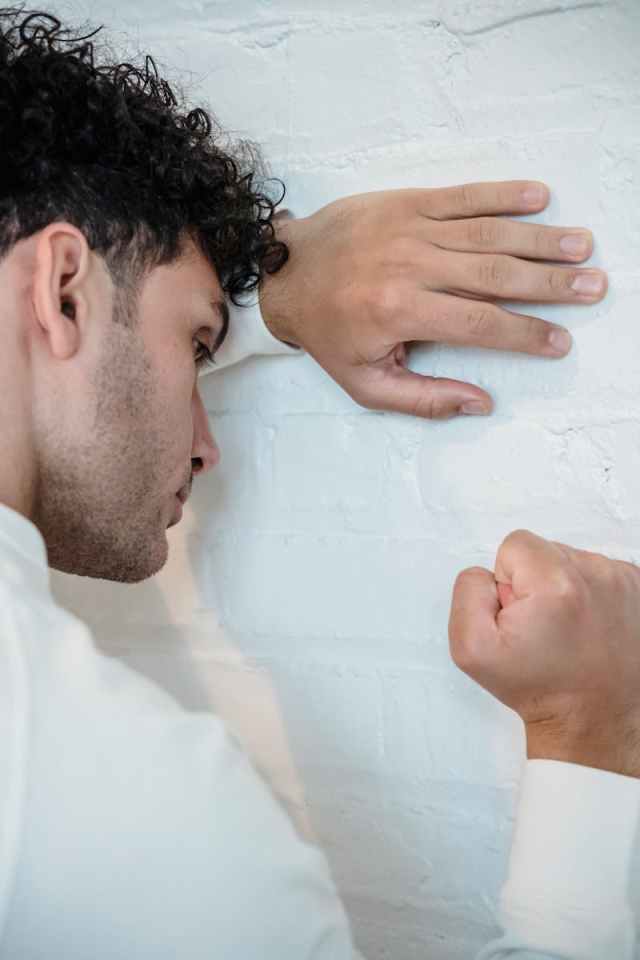 A man with curly hair expresses frustration against a white brick wall in a contemplative pose.