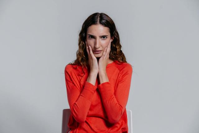 Adult woman sitting indoors with a contemplative facial expression, wearing a red sweater.