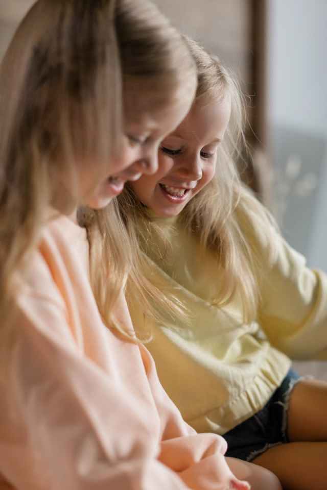 Two smiling blonde sisters enjoying a happy moment indoors, showcasing sibling bonding.