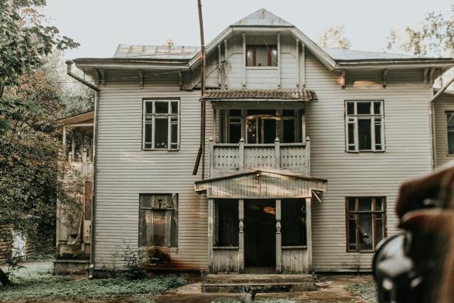 A deserted two-story wooden house with worn out windows and a rustic appearance in a forested area.