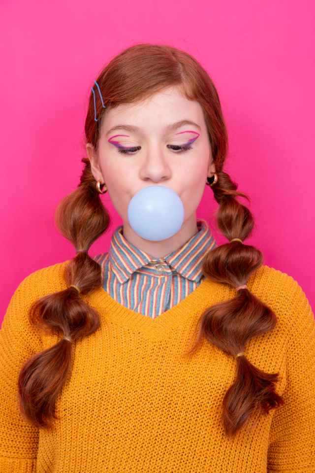 Young redhead girl with braids blowing a bubblegum balloon against a pink studio background.