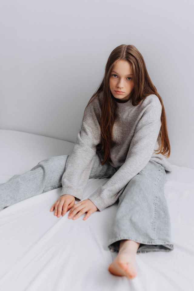 Teenage girl sitting on bed in jeans and sweatshirt, portraying a thoughtful mood indoors.