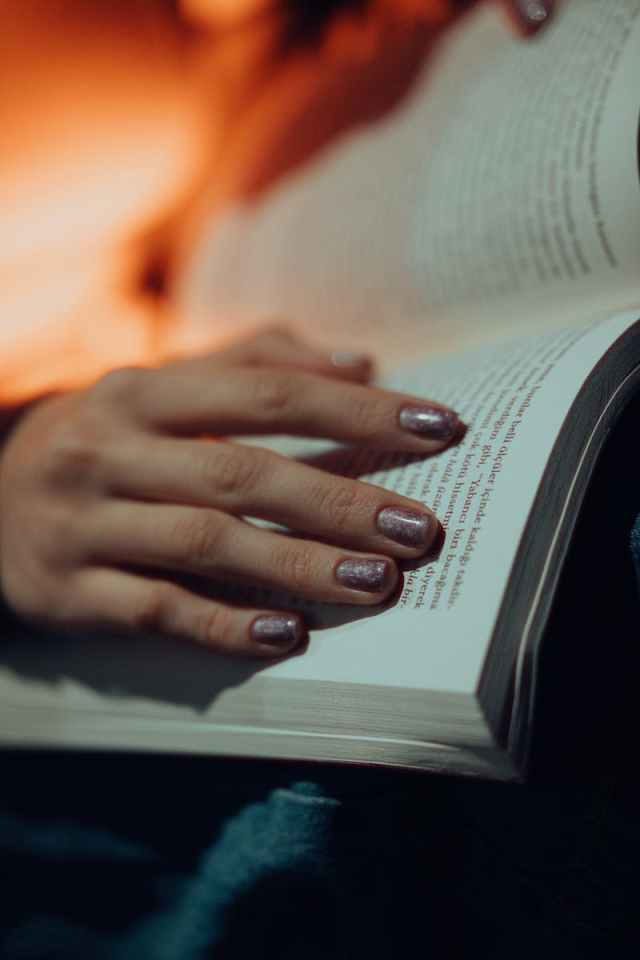 A close-up shot of a woman's hand with glittery manicure holding an open book in dim light.