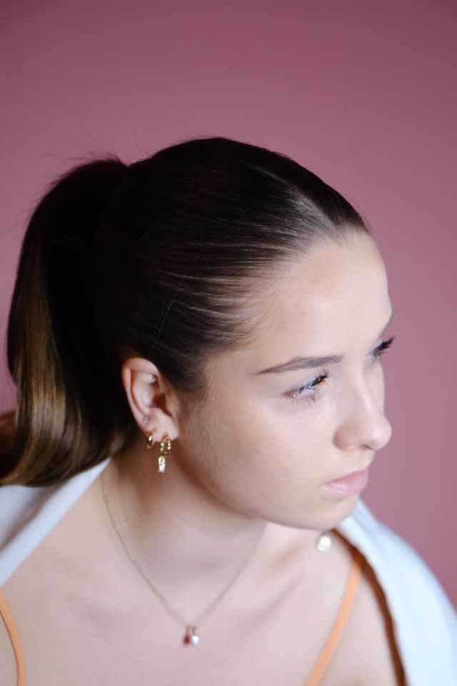 A contemplative woman with brown hair, wearing a necklace and earrings, against a red background.