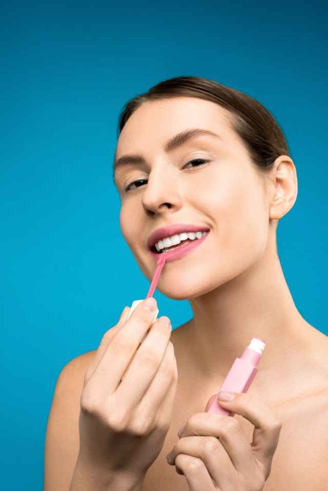 Close-up of a smiling woman applying pink lip gloss, showcasing beauty and makeup techniques.