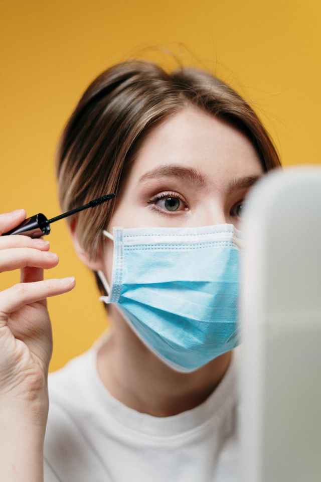 Woman applying mascara with a surgical mask on, reflecting pandemic beauty routines.