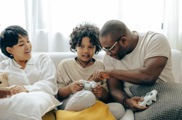 Concentrated black father pointing on joystick buttons explaining rules of playing to son while positive mother watching process