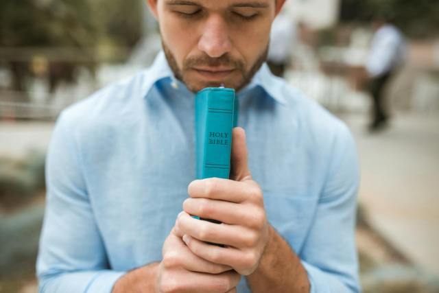 Young man in blue shirt holding a Holy Bible while praying outdoors.
