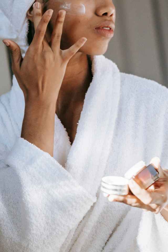 Crop unrecognizable young female in white bathrobe applying moisturizing cream on face after shower
