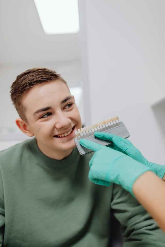Smiling teenage boy at dental checkup with shade guide in hands of dentist wearing green gloves.