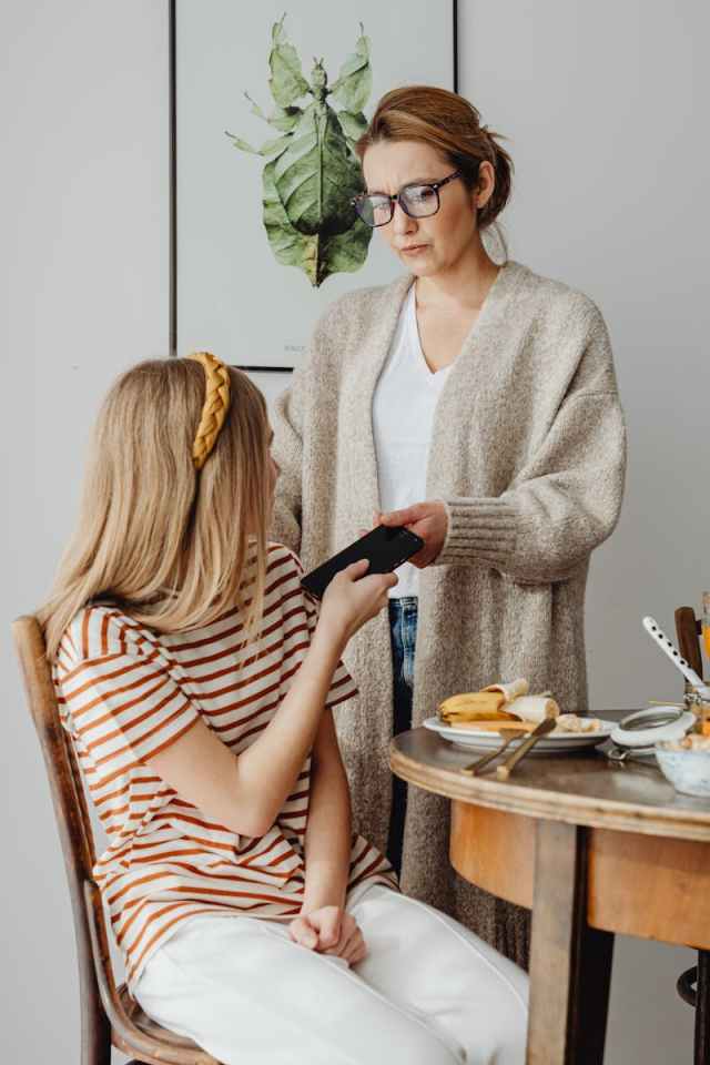 A mother confiscates her teenager's smartphone during breakfast, showing concern.