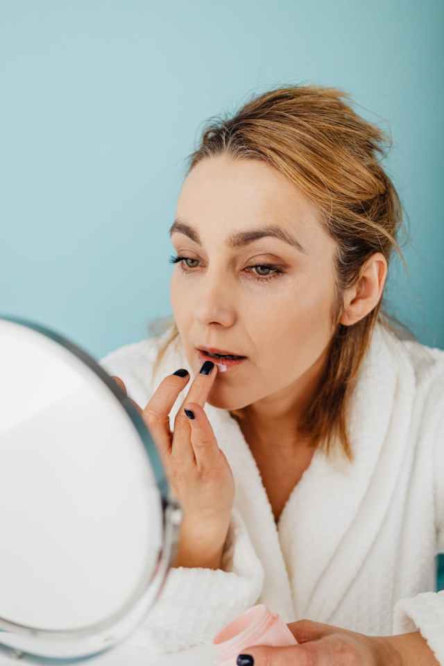 Woman in bathrobe applying lip balm while looking in mirror, focusing on facial care indoors.