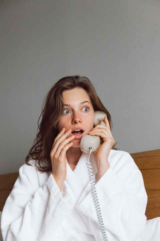 A woman in a bathrobe looks shocked while talking on the telephone indoors.