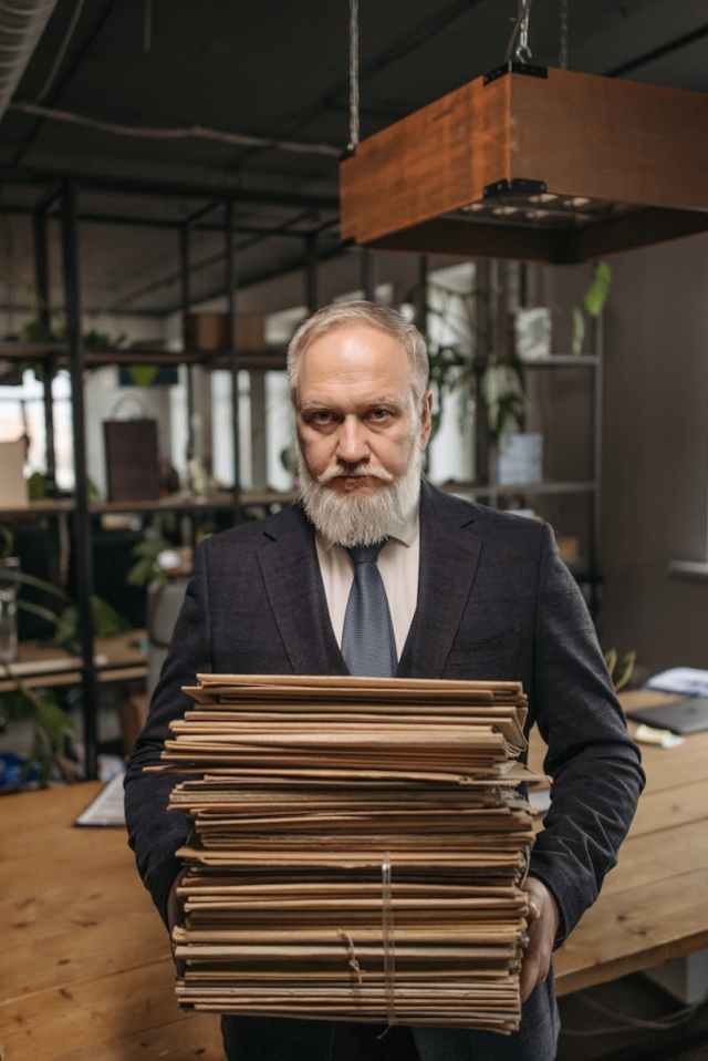 Elderly businessman in a suit holding a stack of papers in a modern office setting.