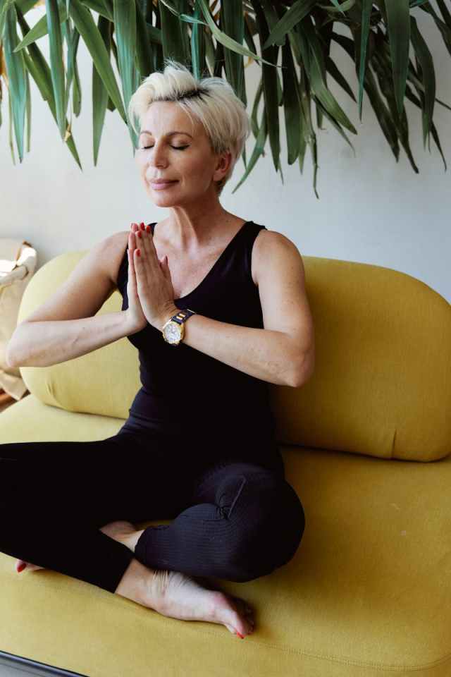 Senior woman meditating on a yellow sofa, embodying calmness and serenity during daytime indoors.