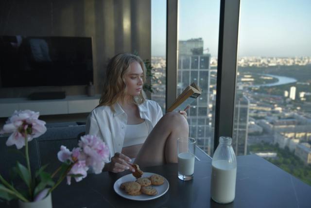 Relaxing scene of a woman reading by a city view, enjoying cookies and milk.