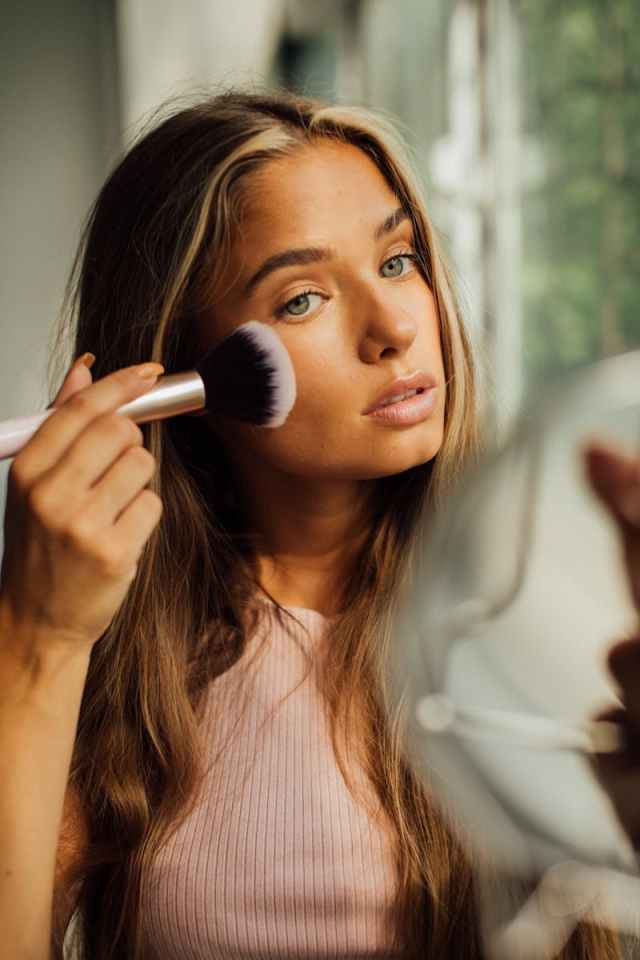 A young woman applies blush using a brush by a window, highlighting natural beauty.