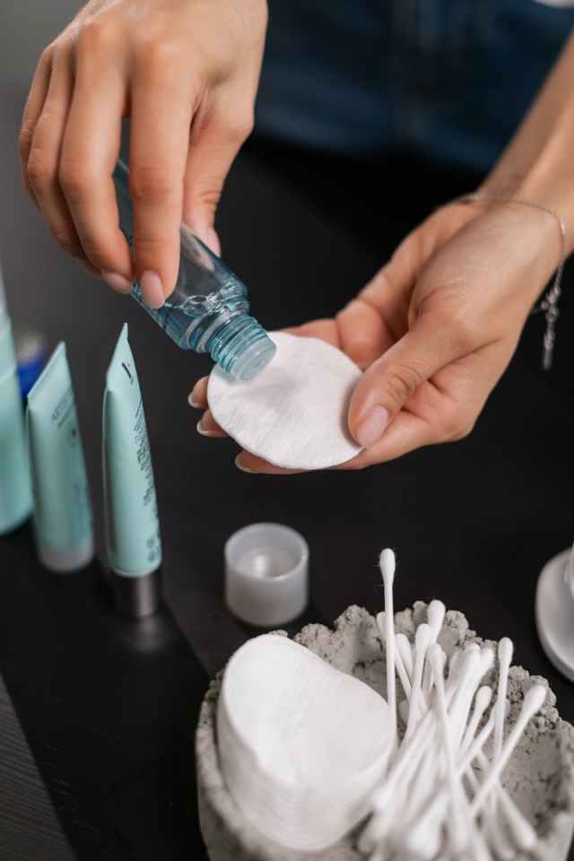 Close-up of hands applying toner to a cotton pad with skincare products in the background.