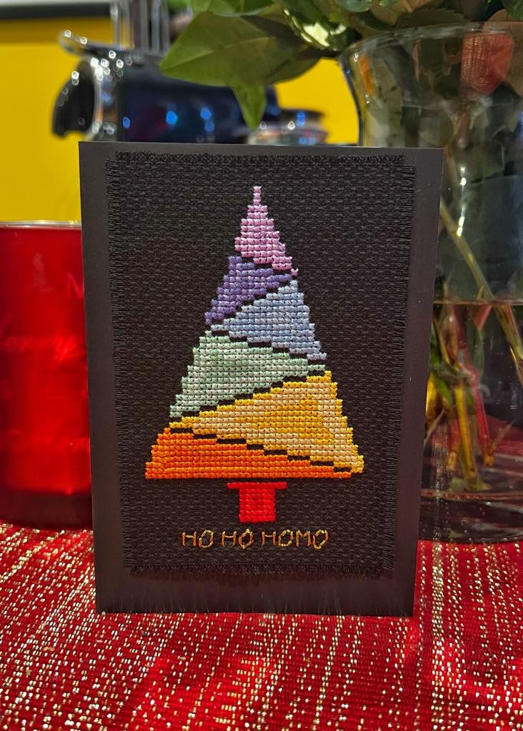 Completed cross-stitch of a Christmas tree made of colourful triangles and the saying "Ho Ho Homo" underneath