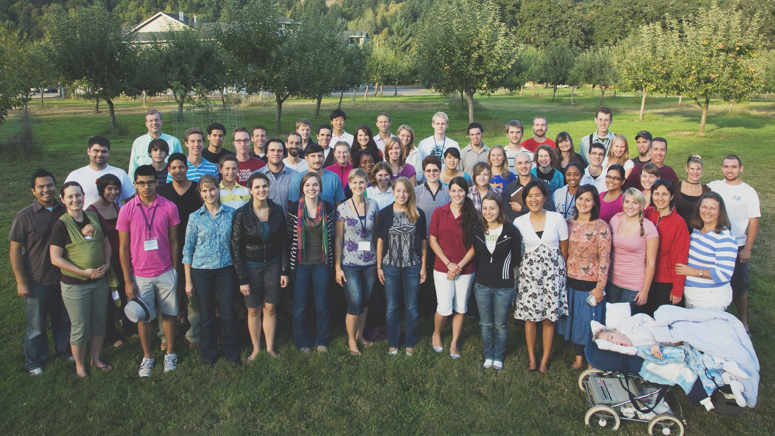 A large, diverse group of people stand together outdoors on a grassy field. They are smiling and casually dressed, with trees and a road in the background. A pile of blankets and a stroller are on the right side of the image. The atmosphere appears friendly and relaxed.