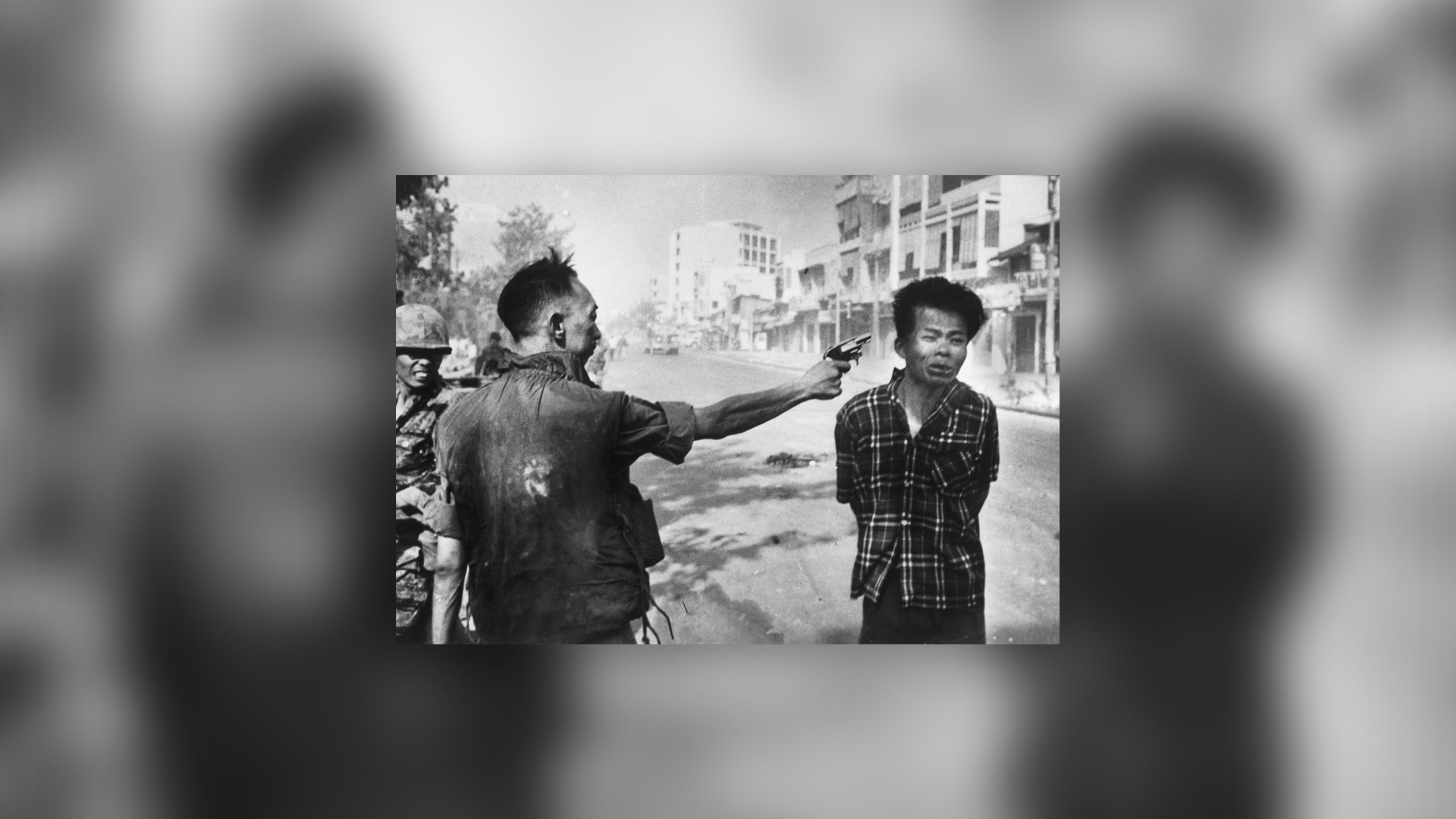 A black-and-white photo shows a man on the left aiming a gun at another man standing to his right. Other individuals are visible in the background while the scene unfolds on a street with buildings. The expressions and stances convey a moment of intense confrontation.