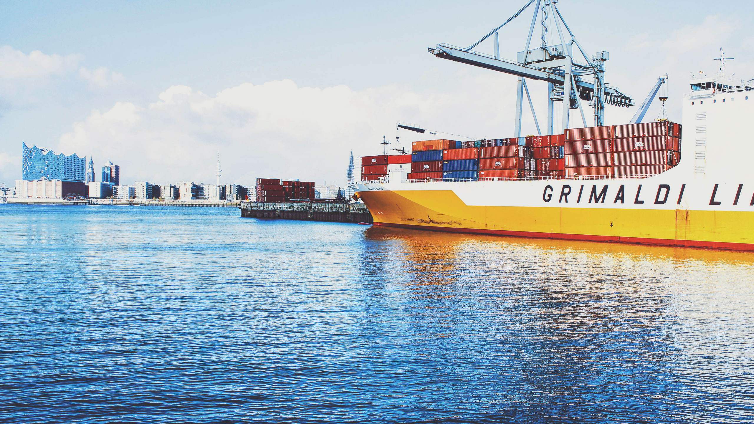 A large cargo ship labeled "Grimaldi Lines" is docked at a port under a clear blue sky. The ship's deck is stacked with multiple colored shipping containers. In the background, there's a cityscape with modern buildings and a prominent concert hall. The calm water reflects the scene.