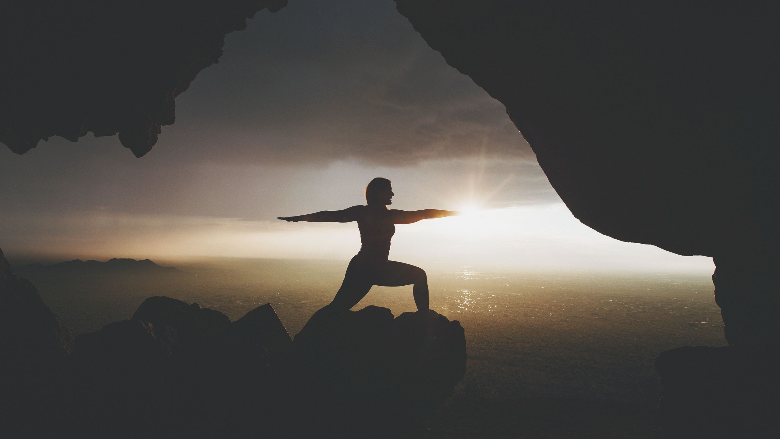 A person is silhouetted in a warrior pose on rocky terrain, situated at the entrance of a cave. The sun is setting in the background, casting a warm glow over the landscape, with a vast expanse of land and distant mountains visible.